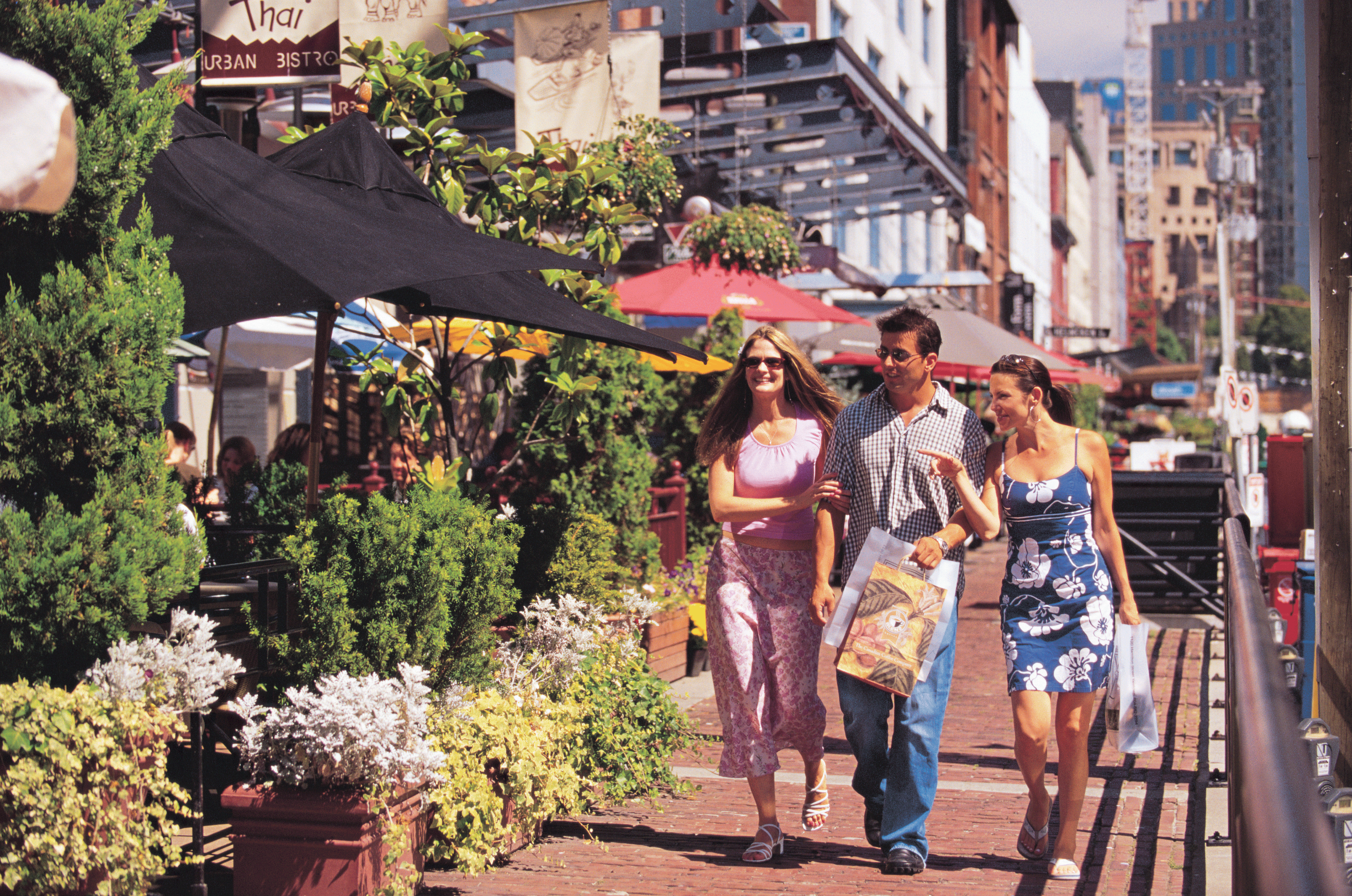 A group of three friends walking down the brick path in Yaletown, with umbrellas and plants on the left side where the patios are. The two women are arm in arm with the man who is holding shopping bags. It is sunny out.