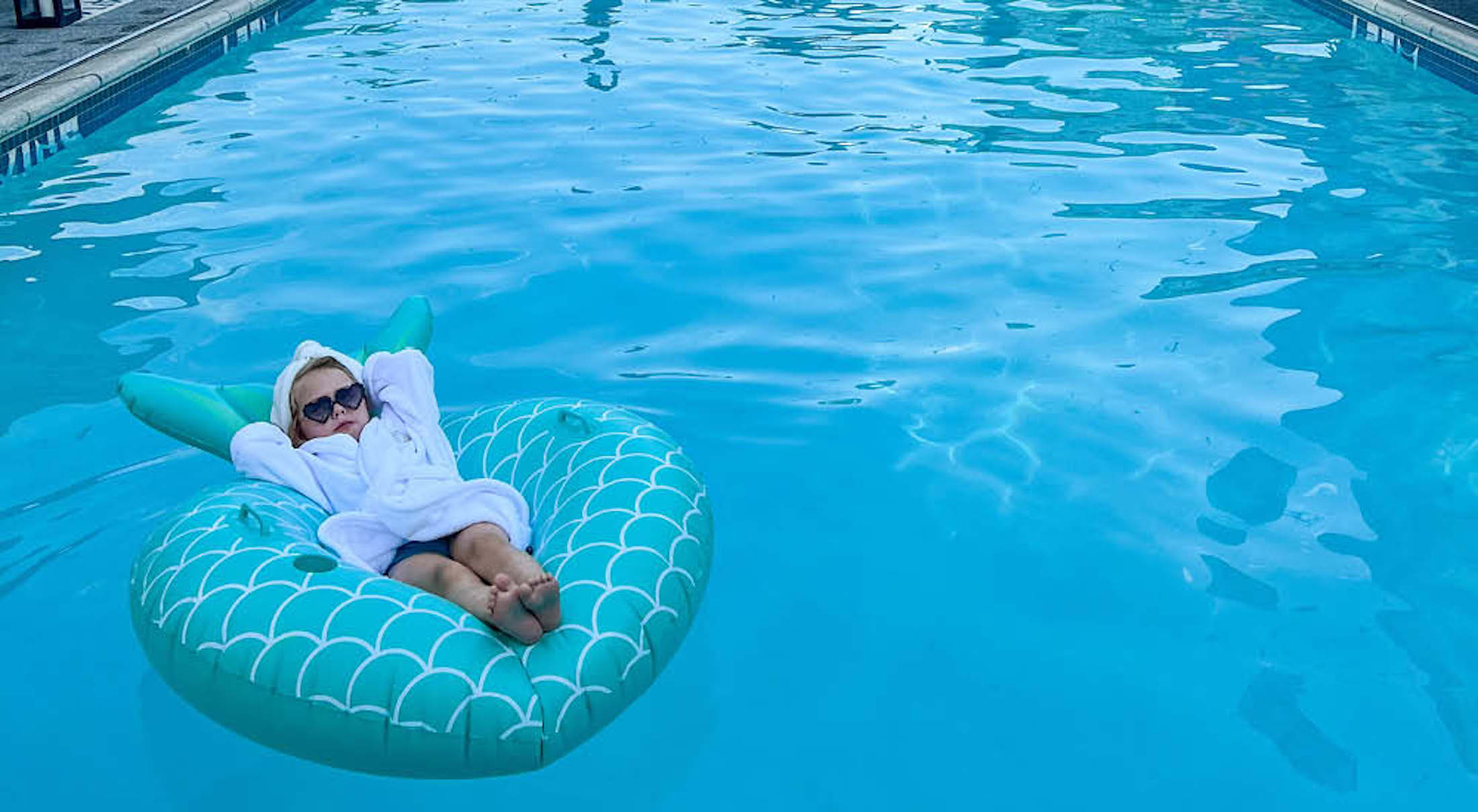 A little girl in a bathrobe relaxing on a pool floatie in an outdoor swimming pool.