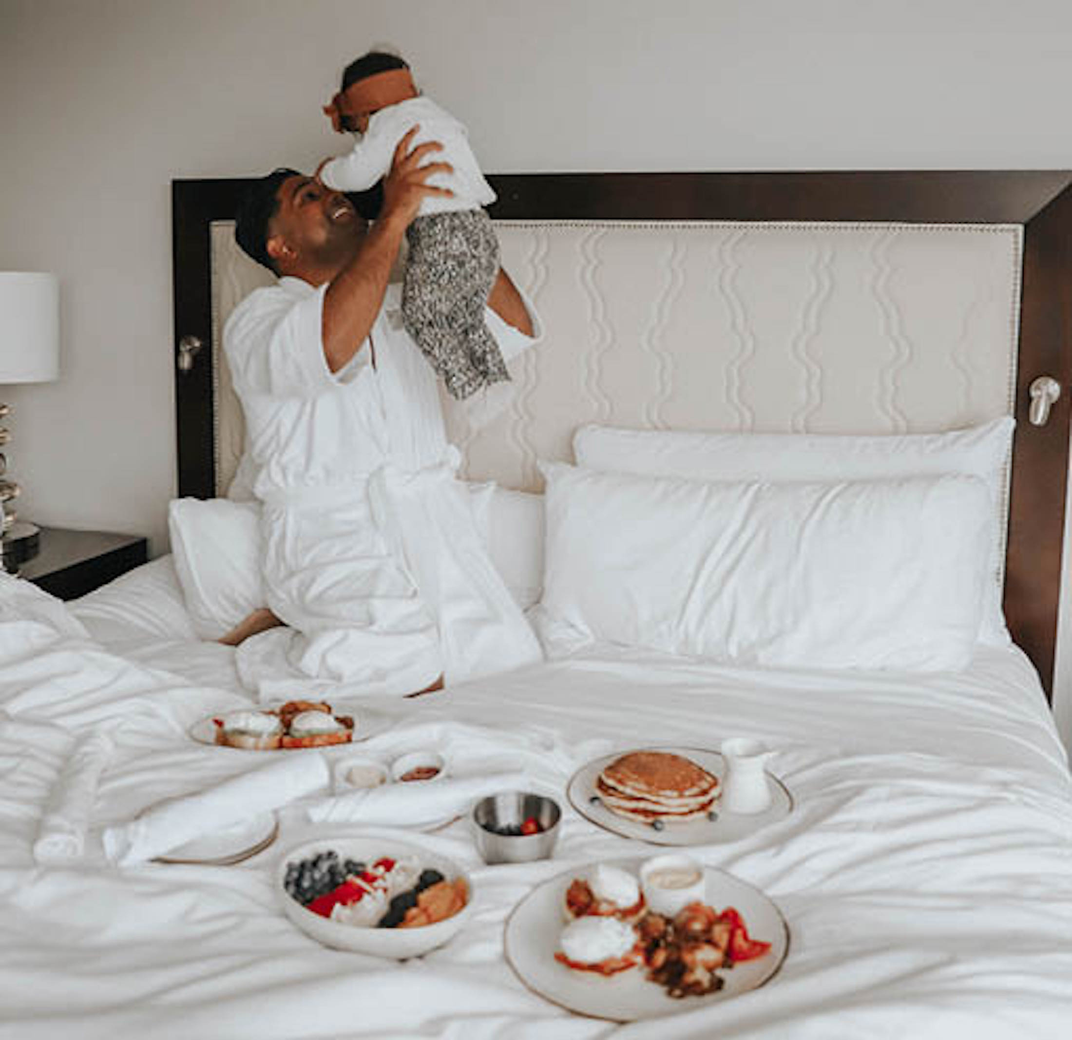 A parent in a bathrobe playing with a toddler on a hotel bed with breakfast