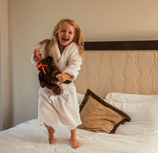 A little girl in a white bathrobe standing on a hotel bed holding a teddy bear.