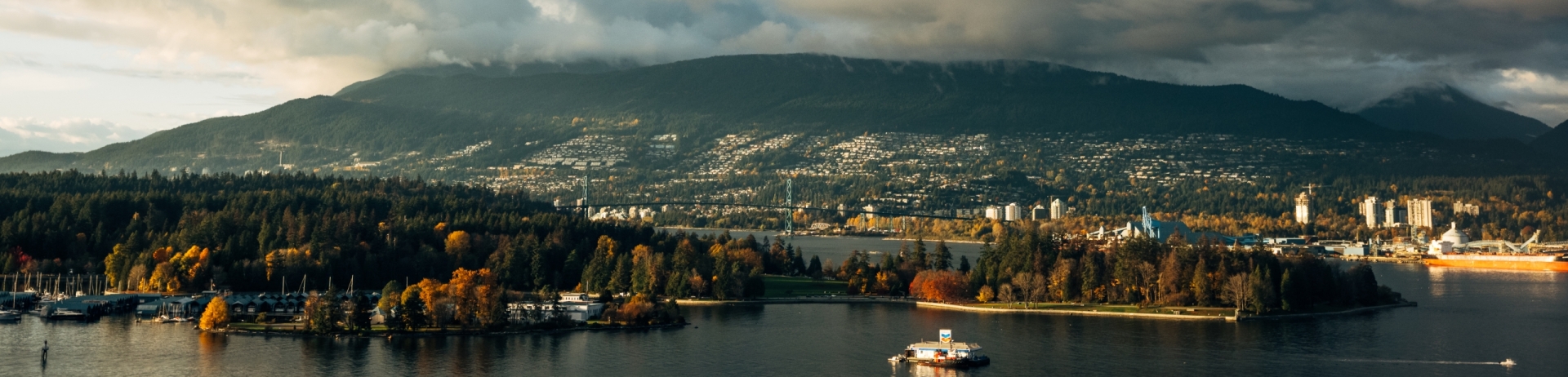 Scenic View of Stanley park in Vancouver, BC taken from Fairmont Waterfront Hotel. Depicts the mountains in the background and the ocean in the foreground. There is sunlight shining through the clouds above which occupy the upper half of the image.