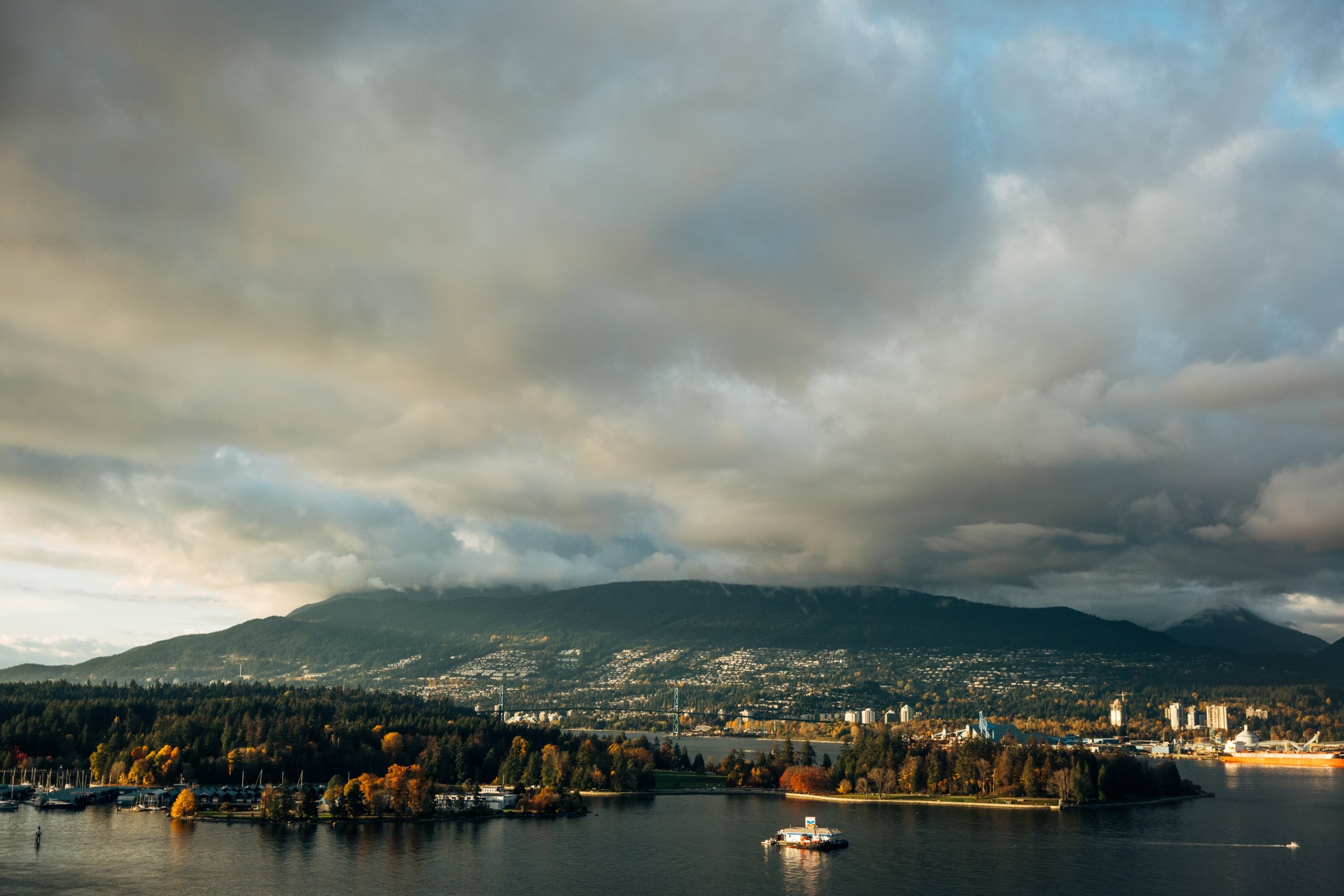 Scenic View of Stanley park in Vancouver, BC taken from Fairmont Waterfront Hotel. Depicts the mountains in the background and the ocean in the foreground. There is sunlight shining through the clouds above which occupy the upper half of the image.