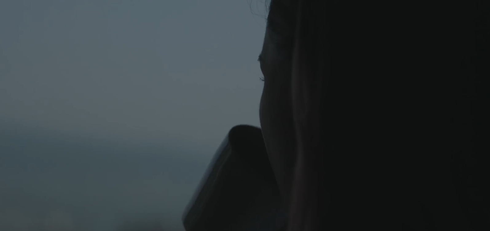 Over-the-shoulder picture of a woman looking out a window while sipping from a mug, the image is primarily in black, blue, and grey hues. It is an extreme close up shot.