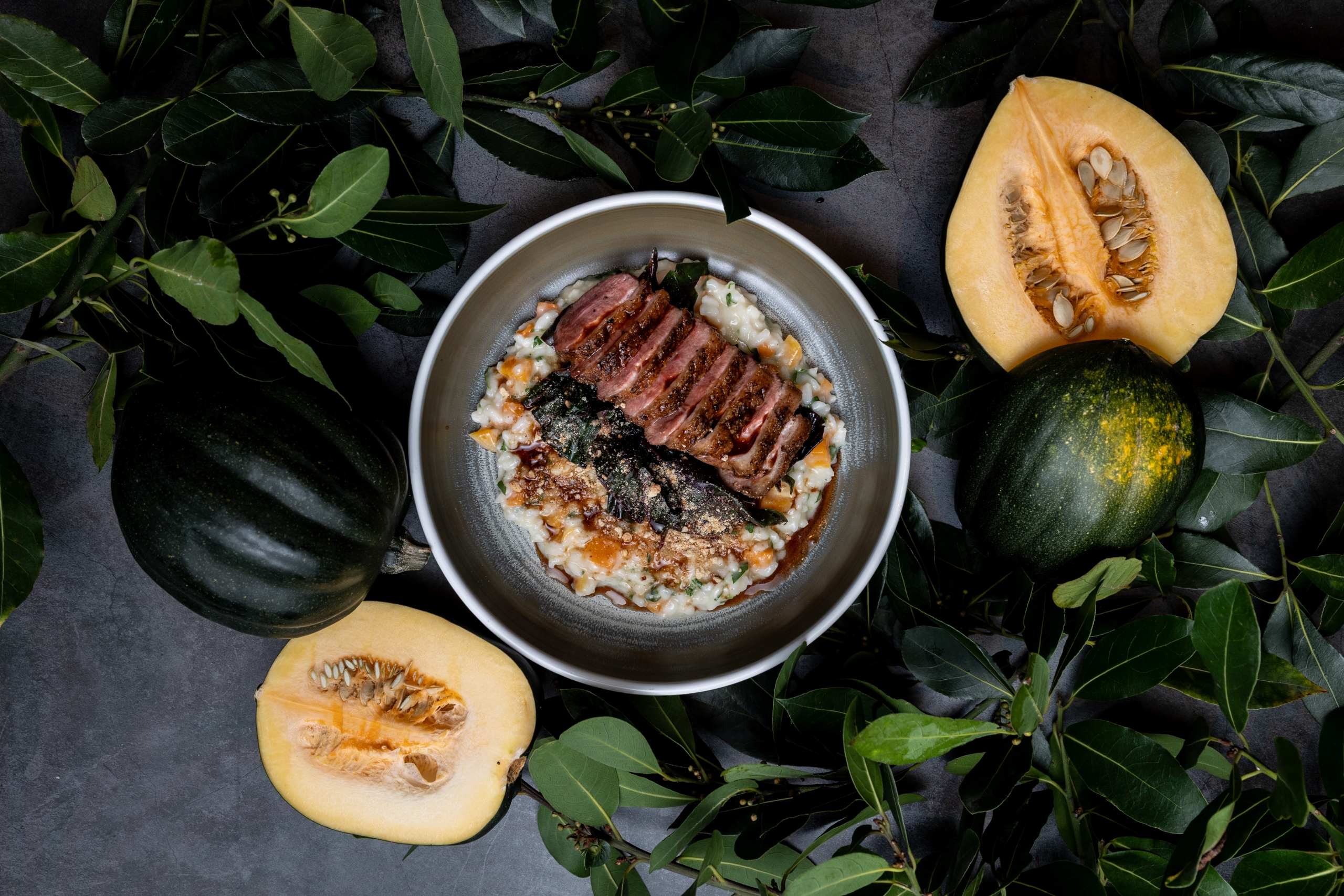 flatlay photo of dish with riotto, duck, and squash with cut squashes and foliage surrounding table