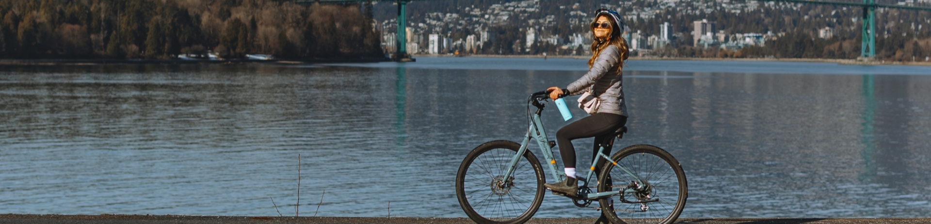 A blonde woman rides a bike with snow on the ground. In the background, you can see beautiful mountains, the harbour, and Stanley Park