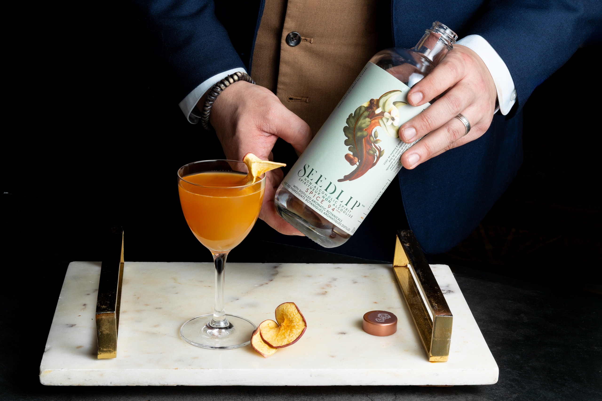 Bartender holding a bottle of Seedlip behind a spirit-free cocktail on a tray.